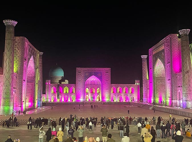 A large historical brown building surrounding a courtyard with many people, lit up with pink lights