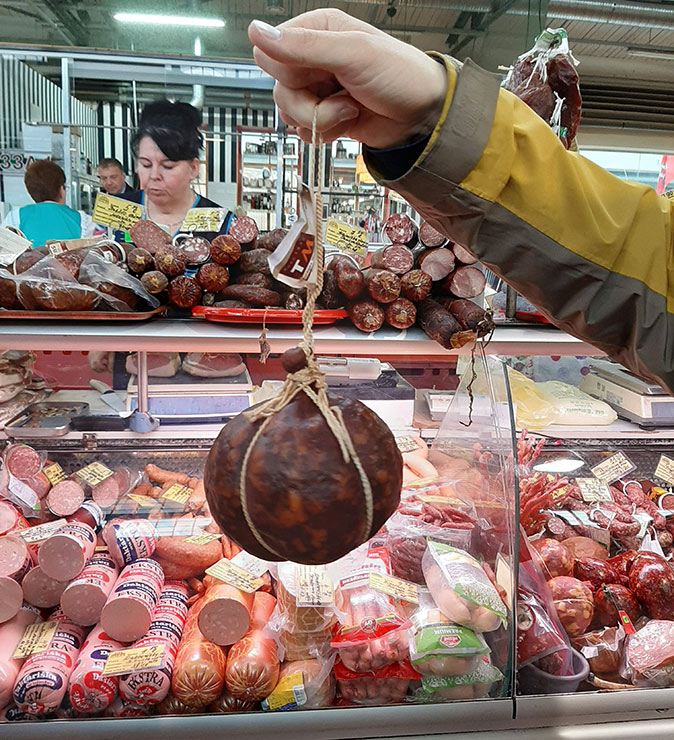 A hand holding a cured sausage in front of a butcher's display