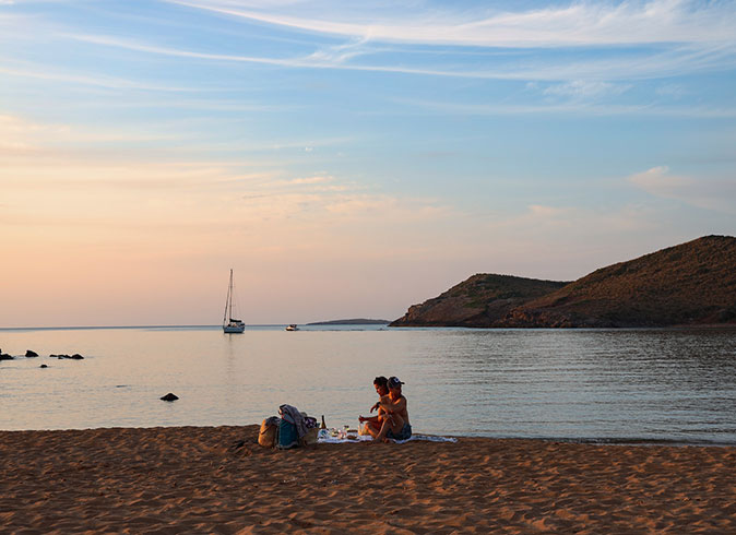 a couple having a picnic on a beach with a sailboat and the sunset in the background