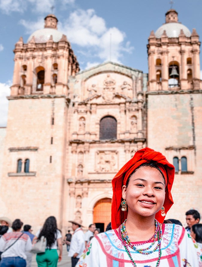 A woman with a bright red scarf and traditional Mexican dress standing in front of a large church