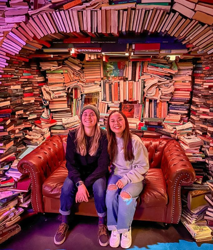A picture of Kristin and her friend sitting on a couch located in an alcove made out of books carefully stacked at Meow Wolf Denver.