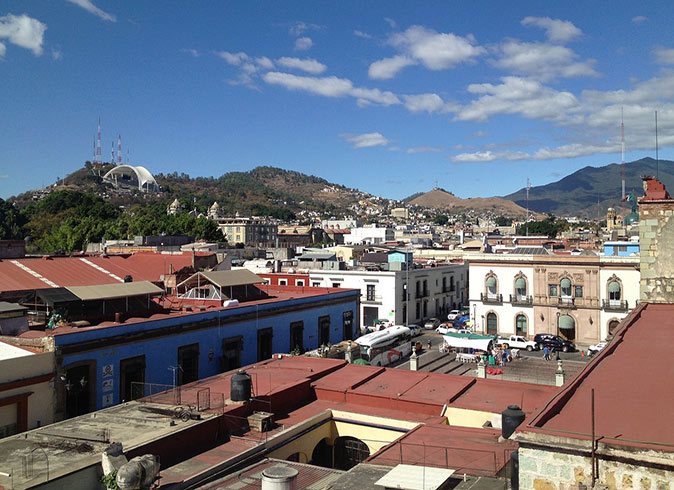 An aerial view of Oaxaca, with many houses with red roofs, a town square and green hills in the background