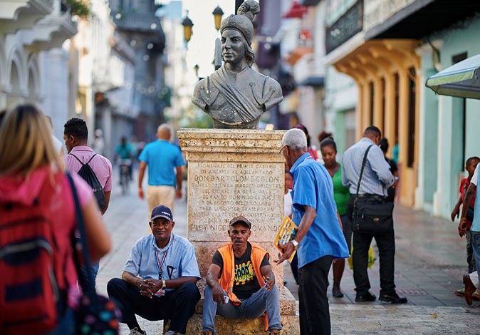 2 people sitting in front of a statue in a pedestrianized street with others walking past, a typical picture of life in the Dominican Republic