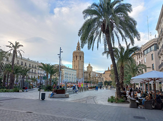 A typical Spanish plaza with palmtrees, several people, terraces and historic buildings