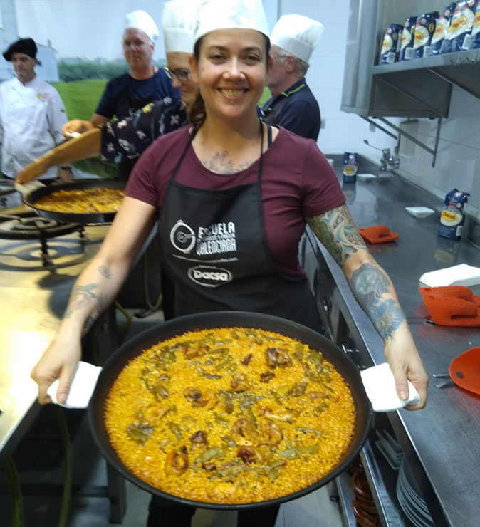A woman smiling, holding a large paella pan with a freshly made Valencian paella