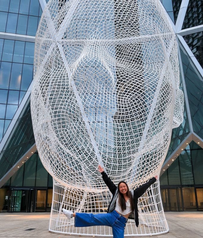 A picture of Kristin posing in front of the Wonderland Sculpture that is located at the entrance to The Bow.