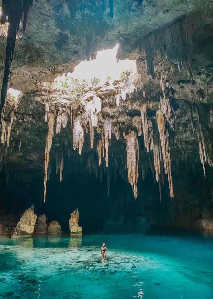 Woman standing in blue water in a large cenote with limestone formations on the ceiling, photo taken at Cenote Xcanahaltun